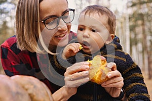 Young woman and her baby son in autumn park, boy playing with helloween pumpkin and eating pumpkin bun.