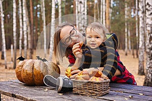 Young woman and her baby son in autumn park, boy playing with helloween pumpkin and eating pumpkin bun.