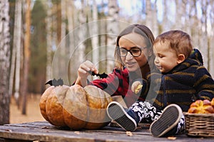 Young woman and her baby son in autumn park, boy playing with helloween pumpkin and eating pumpkin bun.