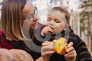 Young woman and her baby son in autumn park, boy playing with helloween pumpkin and eating pumpkin bun.