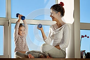 Young woman with her child holding a dumbbell and doing sports. Her daughter with a proud expression lifts the dumbbell