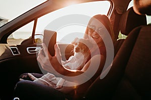 young woman and her cute dog in a car at sunset. travel concept. woman taking a selfie with mobile phone