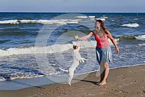 Young woman and her cute small jack russell terrier playing at the beach with a tennis ball. Summer and holidays concept