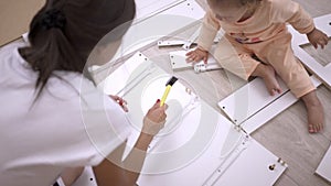 A young woman with her daughter, assemble furniture according to the instructions, hammering nails with a hammer