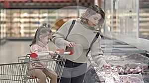 Young woman with her little daughter near showcases with meat in a supermarket. Child sits in the grocery cart and helps