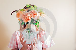 Young woman hides face behind vase with bouquet of flowers. Arrangement with orange roses. Aesthetics in home decor