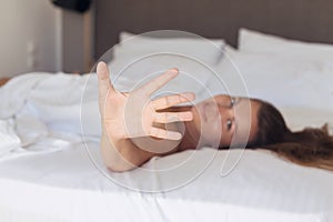 Young woman hides her face from the camera shy and smiling  lying on a bed in the bedroom and covered sheet.