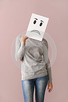 Young woman hiding face behind sheet of paper with drawn emoticon on color background