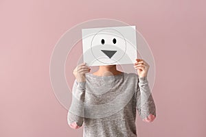 Young woman hiding face behind sheet of paper with drawn emoticon on color background