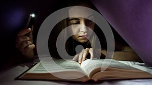 Young woman hiding under blanket and enrapt reading interesting book at nighttime. Girl lighting with the phone as a