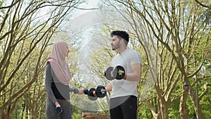 Young woman in hijab and handsome muslim man standing together at green park and training arms using dumbbells. Happy