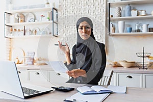 Young woman in hijab Muslim freelancer businesswoman working online at home with laptop and documents. He looks sadly at