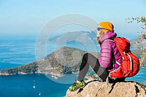 Young woman hiker in bright clothes using digital tablet sit at seaside cliff edge and enjoy the view at mountain peak