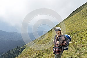 Young woman hiker in cap and sunglasses with large hiking backpack looking at mountain view of the Aibga ridge of the Caucasus