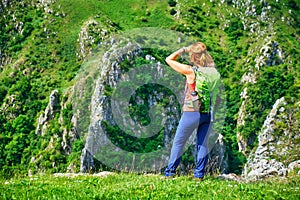 Young woman hiker with a green backpack stands on a grass ridge and looks towards the massive rock walls in Tureni gorge.