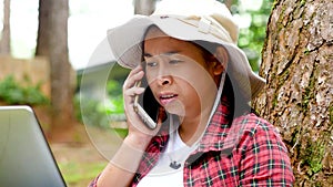 Young woman hikers speak talk on smartphone with a laptop computer on her lap while sitting under a big tree in the park. Lifestyl