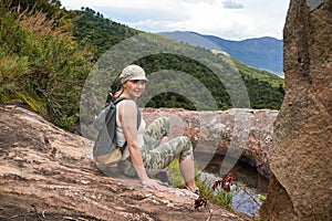 Young woman in hiking clothes, resting on stones near natural water pool, blurred trees and mountains background - typical scenery