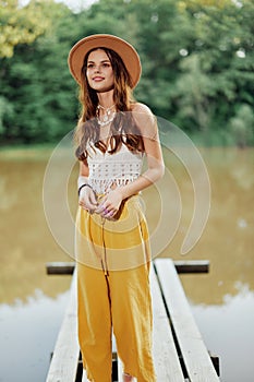 A young woman in a hippie look and eco clothing travels outdoors by the lake wearing a hat and yellow pants in the fall