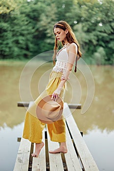 A young woman in a hippie look and eco-dress dancing outdoors by the lake wearing a hat and yellow pants in the summer