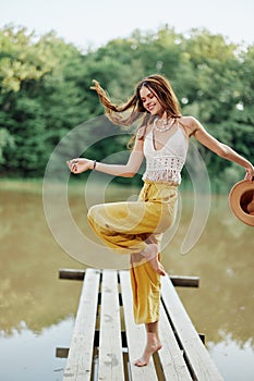 A young woman in a hippie look and eco-dress dancing outdoors by the lake wearing a hat and yellow pants in the summer