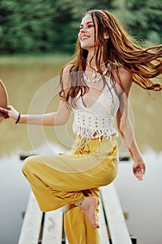 A young woman in a hippie look and eco-dress dancing outdoors by the lake wearing a hat and yellow pants in the summer