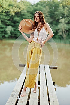 A young woman in a hippie look and eco-dress dancing outdoors by the lake wearing a hat and yellow pants in the summer