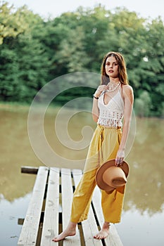 A young woman in a hippie look and eco-dress dancing outdoors by the lake wearing a hat and yellow pants in the summer