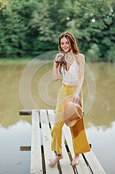 A young woman in a hippie look and eco-dress dancing outdoors by the lake wearing a hat and yellow pants in the summer