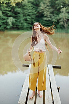 A young woman in a hippie look and eco-dress dancing outdoors by the lake wearing a hat and yellow pants in the summer