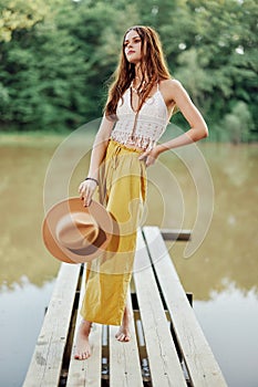 A young woman in a hippie look and eco-dress dancing outdoors by the lake wearing a hat and yellow pants in the summer