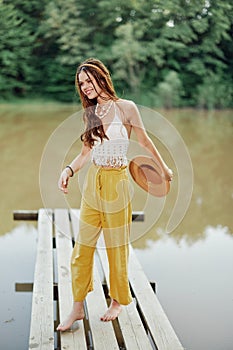 A young woman in a hippie look and eco-dress dancing outdoors by the lake wearing a hat and yellow pants in the summer