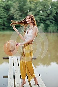 A young woman in a hippie look and eco-dress dancing outdoors by the lake wearing a hat and yellow pants in the summer