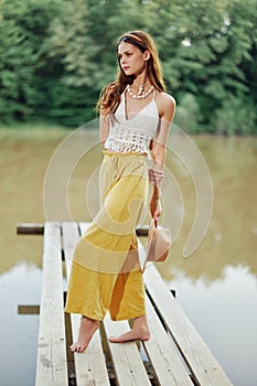 A young woman in a hippie look and eco-dress dancing outdoors by the lake wearing a hat and yellow pants in the summer