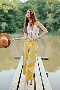 A young woman in a hippie look and eco-dress dancing outdoors by the lake wearing a hat and yellow pants in the summer