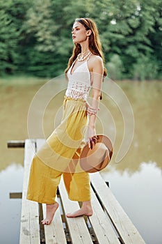 A young woman in a hippie look and eco-dress dancing outdoors by the lake wearing a hat and yellow pants in the summer