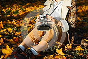 Young woman hipster photographer in boho clothes holding film retro vintage camera while sitting on autumn fallen leaves outdoors