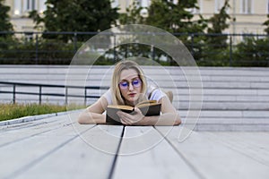 Young woman hipster reading a book lying on the bench in summer