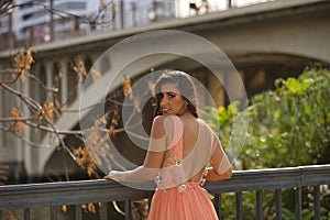 Young woman, Hispanic, beautiful, brunette, wearing an elegant salmon-colored dress, leaning on a railing looking over her