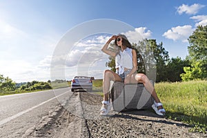 Young woman hitch-hiking on a road with luggage bag at the fields. girl is sitting on baggage and waiting for the car to