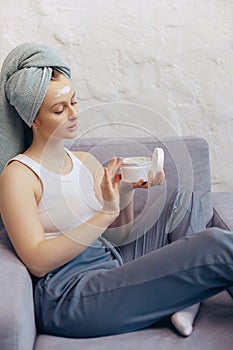 Young woman hold creme jar relaxing on couch, head wrapped in towel