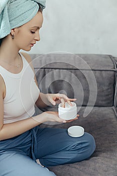 Young woman hold creme jar relaxing on couch, head wrapped in towel