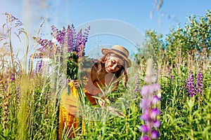 Young woman holding bouquet of lupin flowers walking in summer meadow. Stylish girl picking purple blooms