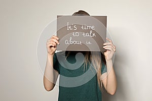 Young woman holding card with words IT`S TIME TO TALK ABOUT IT against background