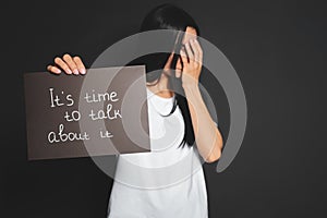 Young woman holding card with words IT`S TIME TO TALK ABOUT IT against dark background