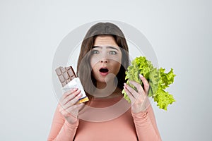Young woman holding chocolate bar in one hand and green salad in another looking surprised standing on isolated white background,