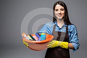 Young woman holding cleaning tools and products in bucket, isolated on grey