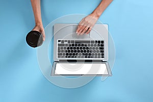 Young woman holding coffee while using laptop on blue background, top view