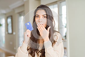 Young woman holding credit card as payment cover mouth with hand shocked with shame for mistake, expression of fear, scared in