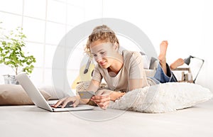 Young woman holding credit card and using laptop computer. Online shopping concept, Lying on living room wooden floor in modern w