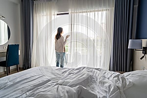 Young woman holding cup of coffee and opening curtains in the morning and looking through the window at home